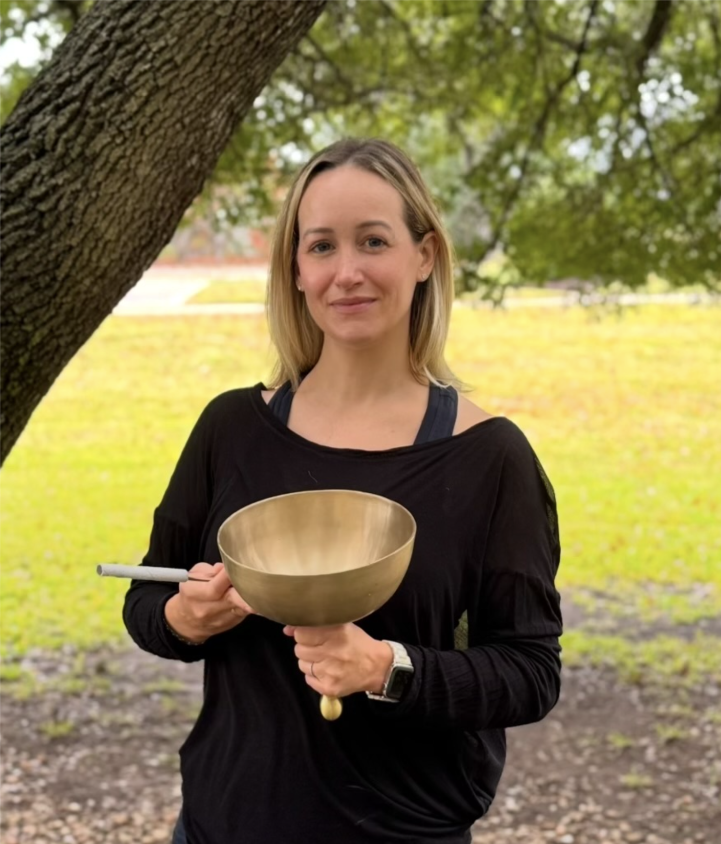 Smiling woman holding a Tibetan singing bowl outdoors A woman in a black top standing outdoors under a tree, holding a large Tibetan singing bowl with a mallet in her hand.