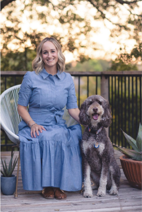 A woman in a blue dress sitting on a chair next to her happy brown and white dog on a wooden deck with potted plants and trees in the background.